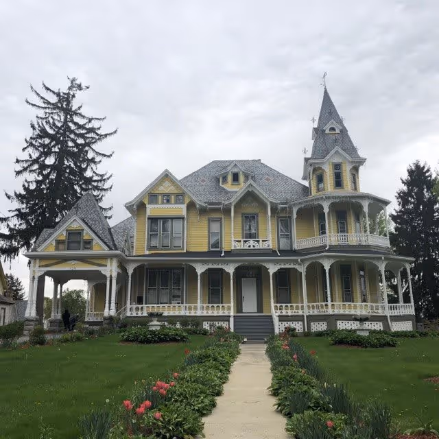 A large, yellow Victorian-style house with a steeply pitched roof, multiple gables, and a prominent turret. The house features a wrap-around porch with white decorative trim and columns. A concrete pathway lined with green plants and pink flowers leads up to the front steps. Tall trees are visible in the background under a cloudy sky.