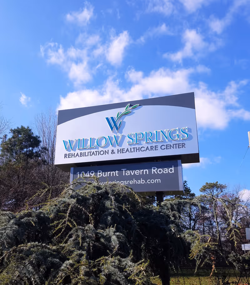 Outdoor view of a large sign for Willow Springs Rehabilitation & Healthcare Center with a clear blue sky and some clouds in the background. The sign includes the address 1049 Burnt Tavern Road and the website willowspringsrehab.com, surrounded by trees and greenery.