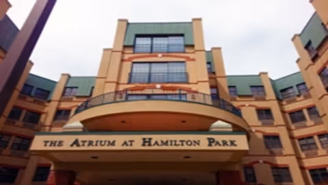 Exterior front view of a multi-story building with a sign that reads 'The Atrium at Hamilton Park'. The building features large windows and a green roof with beige and orange accents.