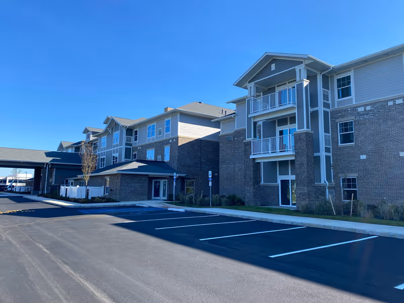 Exterior view of a multi-story retirement community building with balconies, brick and siding facade, adjacent to an empty parking lot under a clear blue sky.