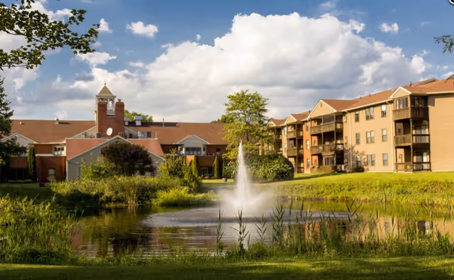Exterior view of a senior living complex with multi-story buildings beside a pond featuring a central fountain under a partly cloudy sky.