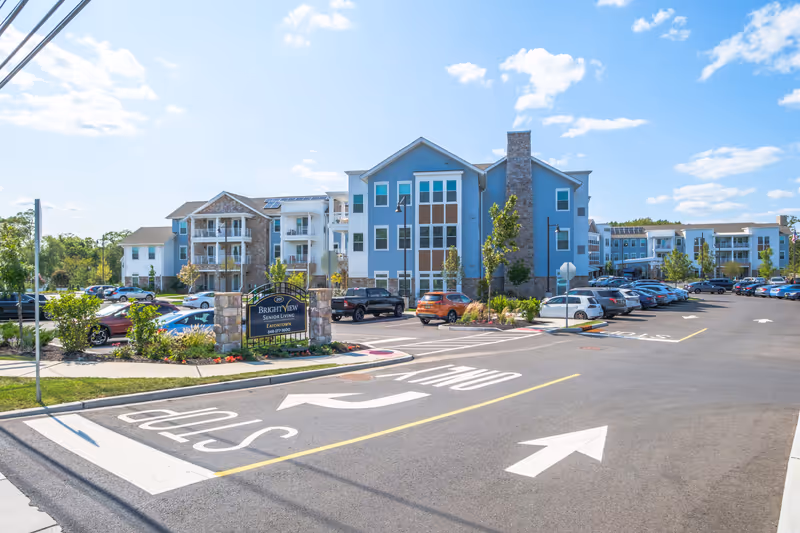 Exterior view of Brightview Eatontown senior living facility showing a large parking lot with multiple cars, a blue and stone building with balconies, and a sign at the entrance that reads 'Brightview Senior Living Eatontown 848-377-3600' under a partly cloudy sky.