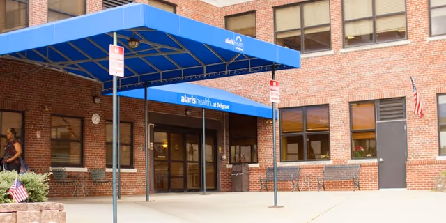 Entrance of Alaris Health at Belgrove facility with a blue canopy over the doorway, brick exterior walls, several windows, benches along the wall, an American flag on the right side, and a person walking near the entrance.