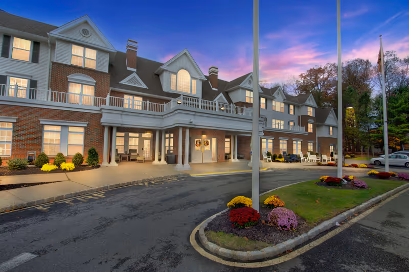 Exterior view of Brighton Gardens of Florham Park senior living facility at dusk, showing a large brick and white-paneled building with multiple windows lit from inside. The entrance has a covered porch with columns, decorated with wreaths on the doors. There are flower beds with colorful flowers and a driveway in front with a 'FIRE' lane marking. Trees and parked cars are visible in the background under a purple and pink sky.