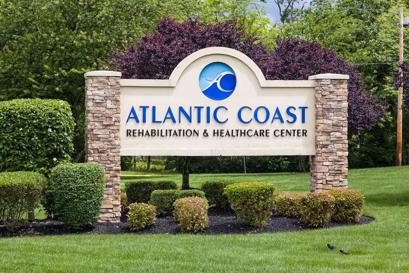 Outdoor sign for Atlantic Coast Rehabilitation & Healthcare Center surrounded by green grass, bushes, and trees.