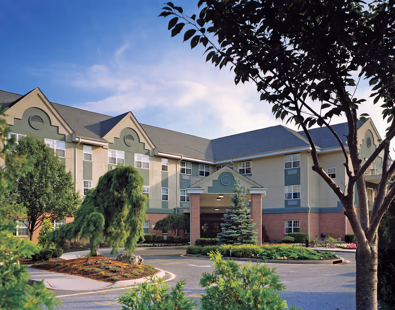 Front exterior of a multi-story senior living facility with a covered entrance, landscaped driveway, and trees.