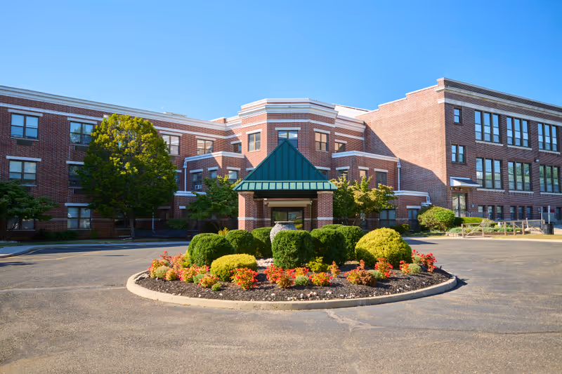 Front entrance of a brick senior living facility with a circular landscaped driveway and a green-roofed canopy.