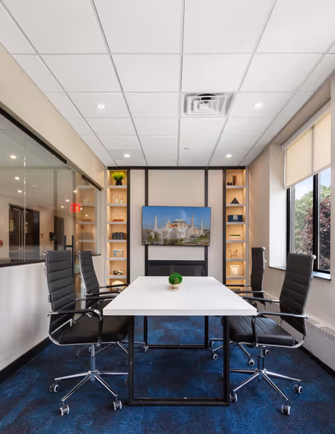 A modern conference room with a white rectangular table surrounded by four black office chairs on wheels. The room has a blue carpet, beige walls, and a ceiling with recessed lighting. A flat-screen TV mounted on the wall displays an image of a historic building. Built-in shelves on either side of the TV hold decorative items and plants. Large windows with roller shades let in natural light.