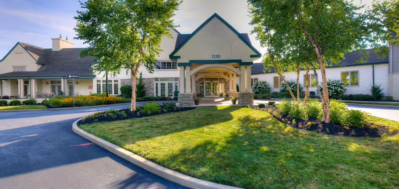 Front exterior of a senior living facility with a covered porte-cochere entrance, landscaped lawn, and circular driveway.
