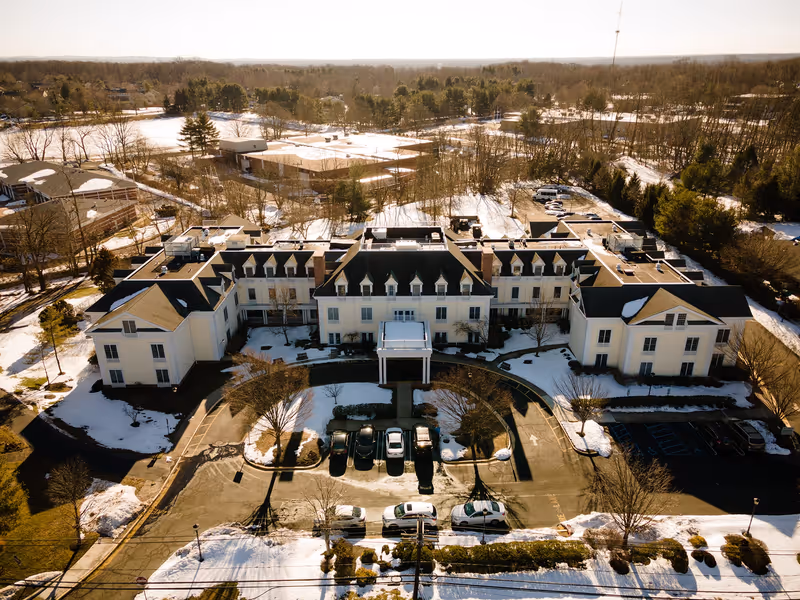 Aerial view of a large, multi-wing building with a dark roof and light-colored walls surrounded by snow-covered ground and trees. Several cars are parked in front of the building, which has a circular driveway and a covered entrance. The surrounding area includes more trees and open land with patches of snow.