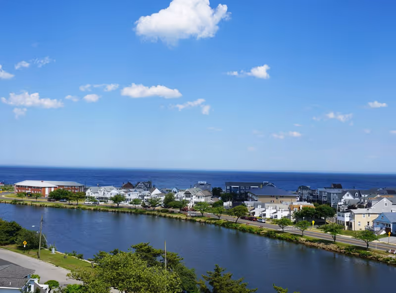 View of a coastal town with houses and buildings along a waterfront, a road lined with trees, and the ocean in the background under a blue sky with scattered clouds.