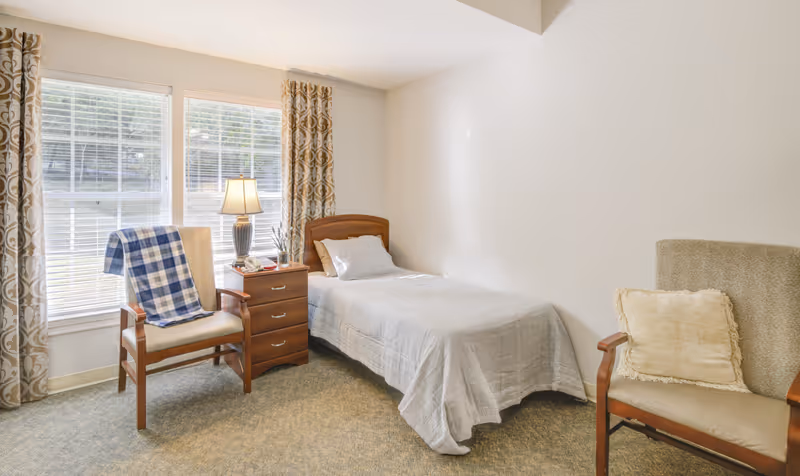 Sunlit bedroom with a single bed, nightstand and lamp, two chairs, and large windows with patterned curtains.