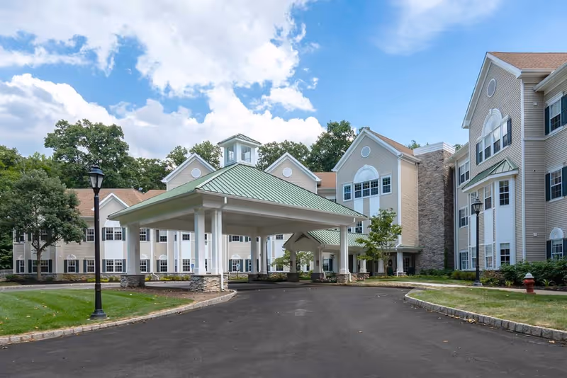 Exterior view of Brandywine Living at Serenade at Princeton showing a large covered entrance with a green roof, surrounded by a multi-story beige building with white trim and multiple windows. The driveway curves around the entrance, and there are trees, grass, and lamp posts in the landscaped area.