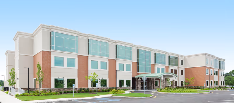 Exterior view of a modern three-story senior living facility building with large windows, a covered entrance, and landscaped greenery around the parking area under a clear blue sky.