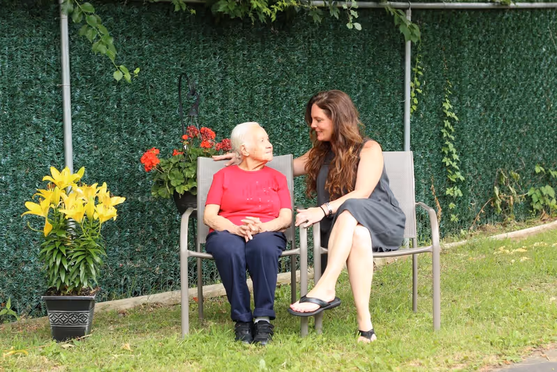 An elderly woman in a red shirt and dark pants sits on a chair outdoors next to a younger woman in a gray dress and black sandals. They are sitting on a grassy area with a green leafy fence behind them and a pot of yellow flowers nearby. The younger woman has her arm around the elderly woman, and they are smiling at each other.