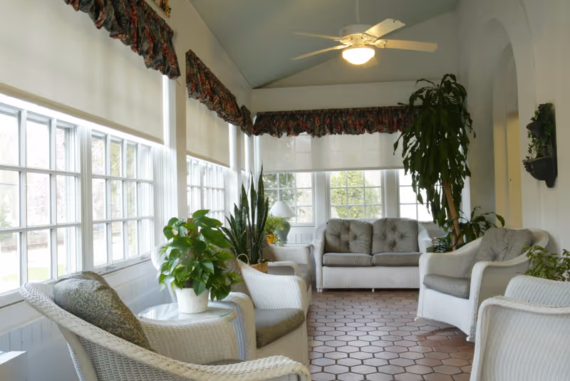 A bright sunroom with large windows covered by white roller shades and floral valances. The room features white wicker furniture with gray cushions, including a sofa and several armchairs arranged around a small glass-top table with a potted plant. There are additional green plants in the corners, a ceiling fan with a light, and terracotta tile flooring.