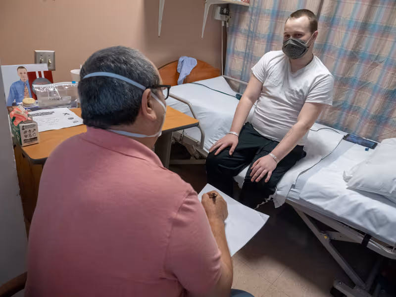 Two masked people in a small care room with a medical bed, bedside table, and curtains.