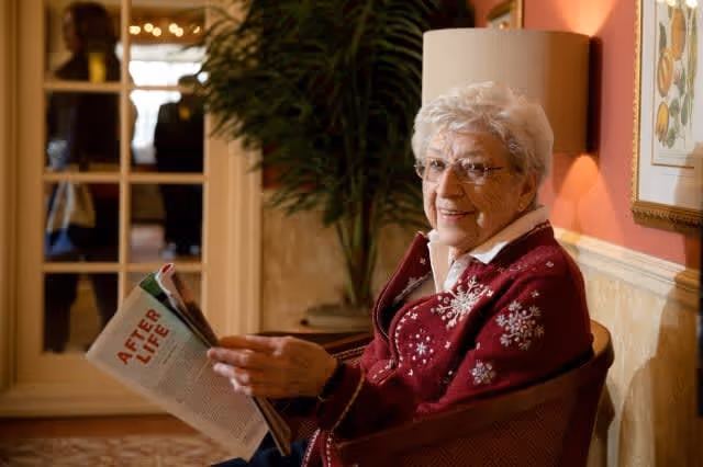 An elderly woman with white hair and glasses is sitting in a chair indoors, wearing a red sweater with white snowflake patterns, and holding a magazine titled 'AFTER LIFE'. There is a plant and framed artwork on the wall behind her.