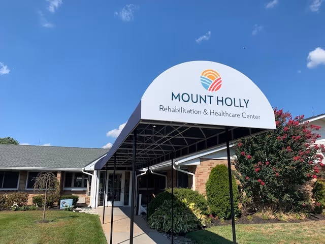 Entrance canopy and sign reading 'Mount Holly Rehabilitation & Healthcare Center' at the front of a single-story brick building with landscaping under a blue sky.