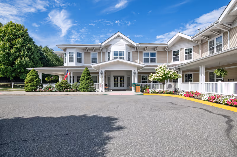 Front entrance of a two-story senior living building with a covered porte-cochère, landscaped flowerbeds, and an American flag.