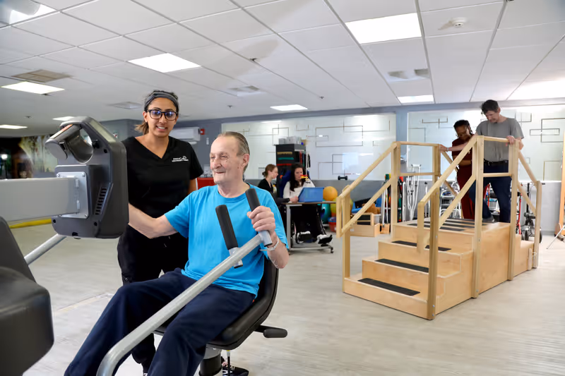 A senior man in a blue shirt exercises on a seated rowing machine with the assistance of a female staff member wearing black scrubs. In the background, another staff member helps a man walk up wooden stairs with handrails in a rehabilitation or physical therapy room. Other staff and therapy equipment are visible in the room.
