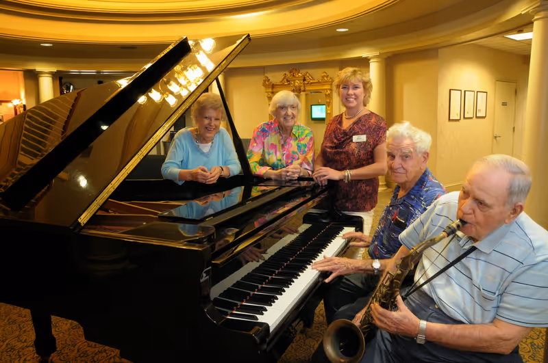 A group of five seniors gathered around a grand piano in a warmly lit room. Two elderly women are seated at the piano, one elderly man is playing the saxophone, another elderly man is seated next to the piano, and a woman standing behind them is smiling. The room has elegant decor with columns and framed pictures on the wall.