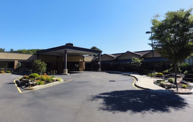 Single-story care center front entrance with a covered drop-off, curved driveway and landscaped beds under a clear blue sky.