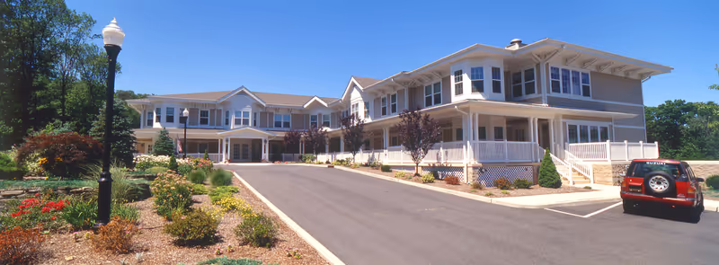 Exterior view of a two-story senior living facility building with a driveway leading to the entrance. The building has multiple windows, a covered porch with white railings, and landscaped gardens with various plants and flowers. A red Suzuki vehicle is parked in the parking lot on the right side. The sky is clear and blue.