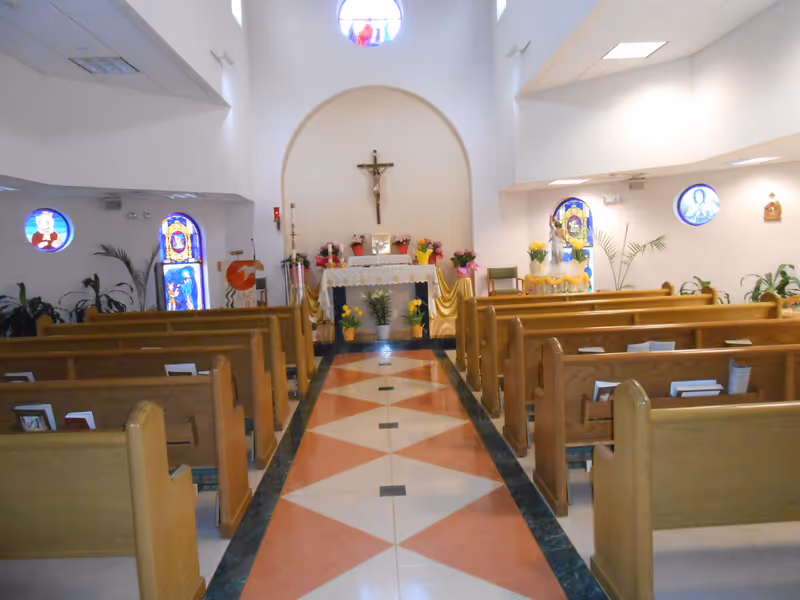 Interior view of a chapel with wooden pews arranged in rows facing an altar. The altar is decorated with flowers, candles, and a crucifix mounted on the wall above it. Stained glass windows are visible on the side walls, and the floor has a geometric pattern in shades of red, white, and green.