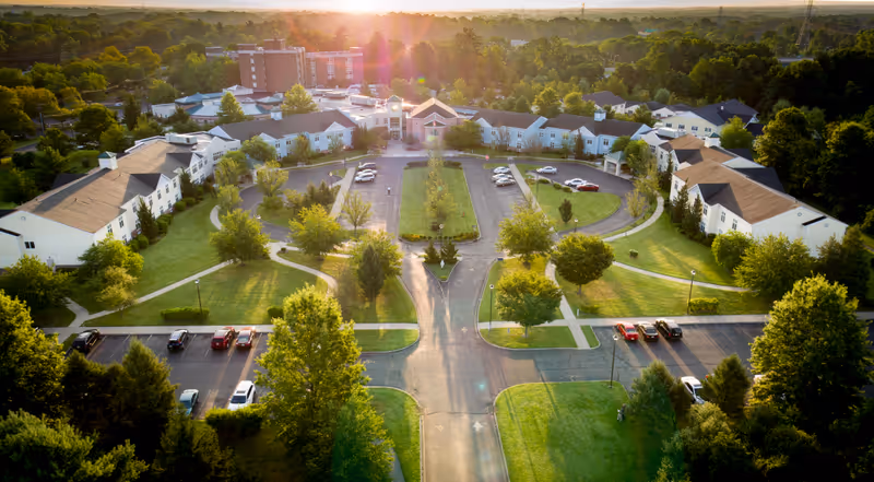 Aerial view of The Terraces at Lawrence senior living facility during sunset, showing multiple white buildings with brown roofs arranged around a central driveway and parking areas, surrounded by green lawns and trees.