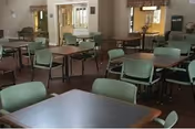 A dining room with multiple square tables and green chairs arranged neatly. The room has beige walls, windows with curtains, and a carpeted floor. There is a water dispenser visible in the background.
