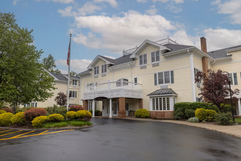 Exterior view of a large, light yellow assisted living facility building with white trim and multiple windows. The building has a covered entrance supported by brick columns and a balcony above it. There are well-maintained bushes and trees around the driveway, and an American flag is visible on a flagpole near the entrance. The sky is partly cloudy.