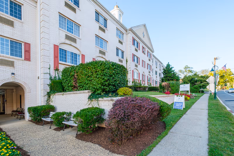 Front exterior of the Mira Vie at Fanwood senior living building with landscaped entrance, benches, and sidewalk signs.