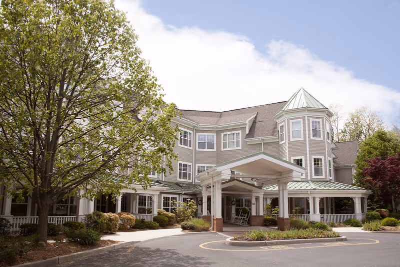 Exterior view of a senior living facility building with a covered entrance, surrounded by trees and landscaping under a partly cloudy sky.