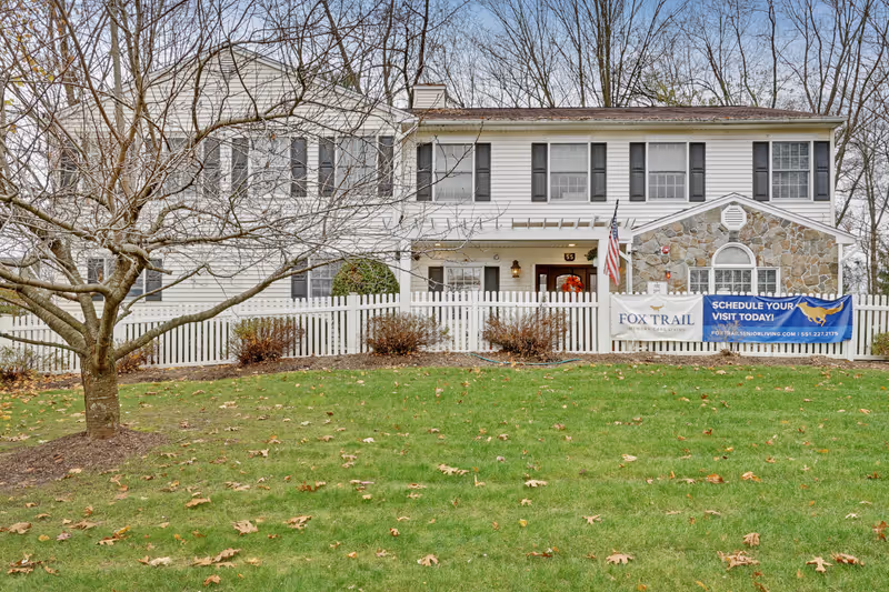 Exterior view of a two-story white building with black shutters and a stone accent wall, surrounded by a white picket fence and a lawn with scattered fallen leaves. A leafless tree stands in the foreground. Two banners hang on the fence, one reading 'FOX TRAIL MEMORY CARE LIVING' and the other promoting scheduling a visit with contact information.