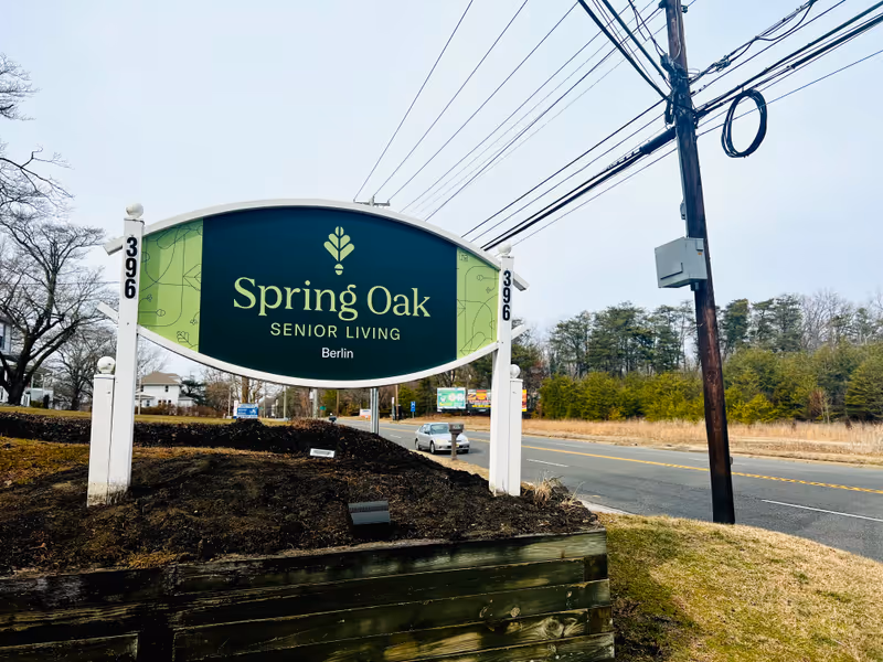 Outdoor view of a large sign for Spring Oak Senior Living in Berlin, positioned on a grassy area next to a road with trees and power lines in the background.