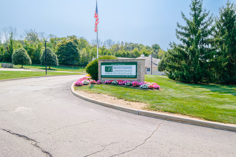 Entrance sign for Bentley Commons at Paragon Village, a senior living community, surrounded by green grass, colorful flowers, and trees, with an American flag on a pole behind the sign.