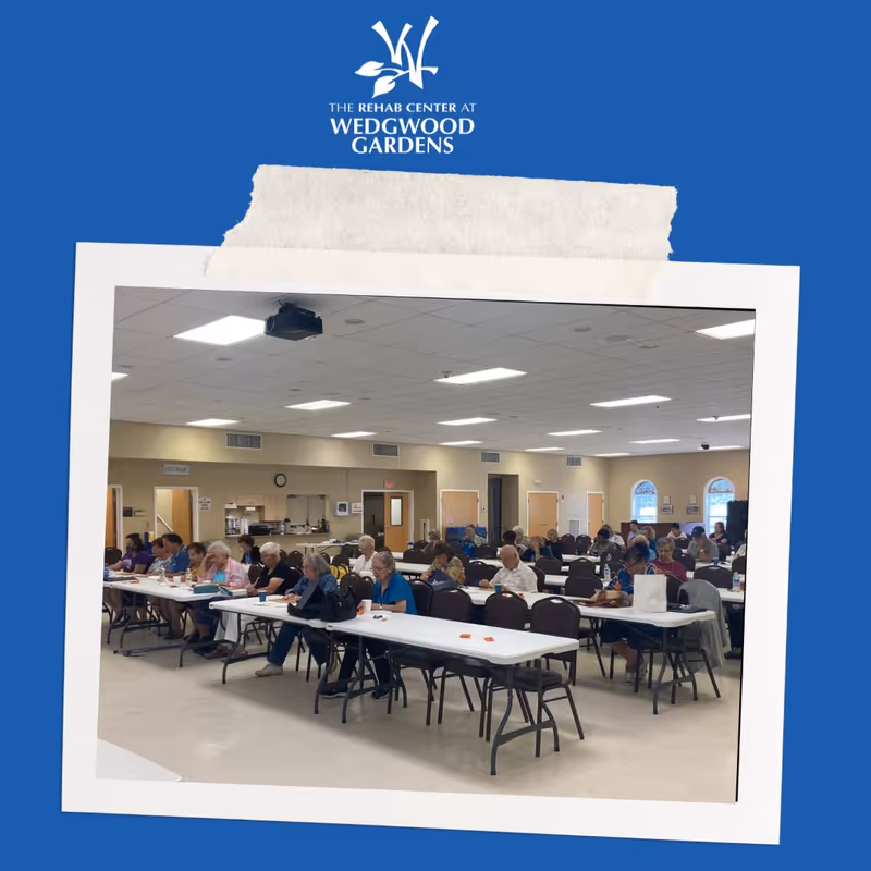 A large room with many elderly people seated at long tables, engaged in activities or writing. The room has a high ceiling with fluorescent lights, beige walls, and several doors and windows. The setting appears to be a community or activity room within a senior living or rehab facility.