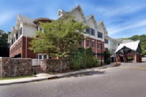 Exterior view of a multi-story senior living facility with a brick and white facade, a covered entrance, and a tree in front under a blue sky.