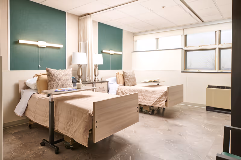 A bright and clean room with two hospital-style beds, each with beige bedding and pillows. Each bed has an overbed table with items on top, and there are two bedside tables with matching lamps between the beds. The room has large windows letting in natural light and green accent panels on the wall behind the beds.