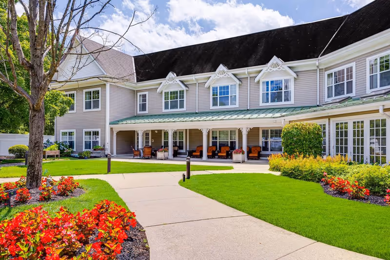 Exterior view of a senior living facility building with beige siding and white trim. The building has two stories with multiple windows and a covered porch area furnished with orange cushioned chairs. The foreground features a well-maintained garden with bright red flowers, green shrubs, a tree, and a paved walkway leading to the entrance. The sky is partly cloudy with blue patches.