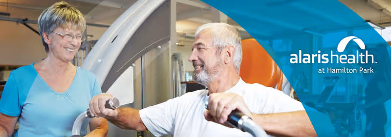 An elderly man using exercise equipment in a fitness or rehabilitation setting, assisted by a smiling female staff member wearing a blue shirt. The background shows gym equipment and a bright, clean environment.