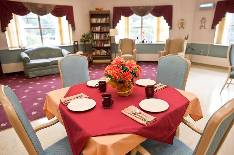 Dining table set for four with a floral centerpiece in a bright senior living common room with upholstered chairs, sofas, and bookshelves.