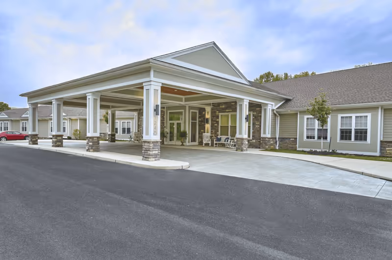Front exterior view of Harmony Village CareOne at Cherry Hill showing a covered entrance with white pillars and stone bases, a driveway, and part of the building with windows and a small tree in front.