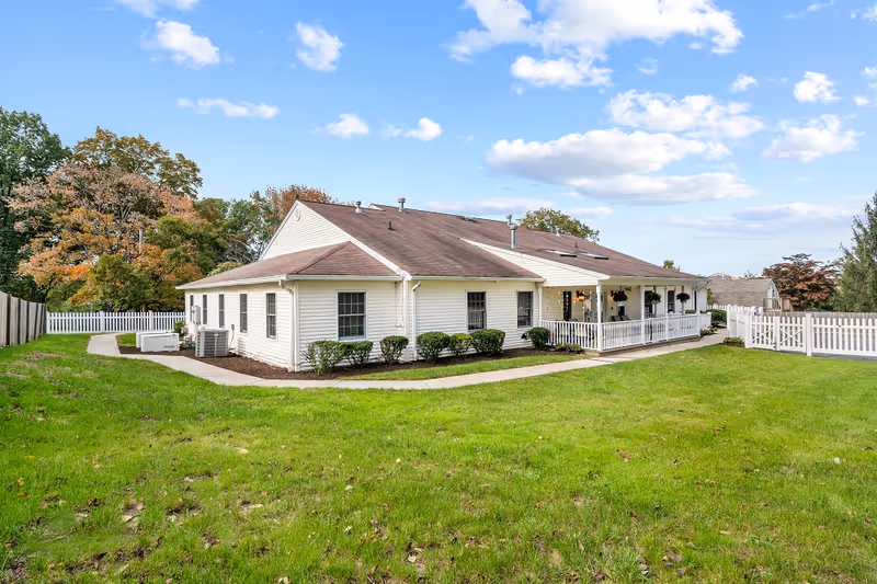 Single-story white building with a brown roof surrounded by a green lawn and white picket fences under a partly cloudy blue sky. There are shrubs along the building and a covered porch with hanging plants.