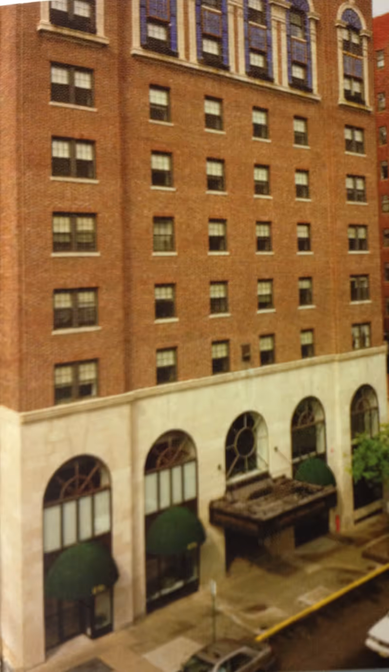 Exterior view of a multi-story brick building with large arched windows on the ground floor, green awnings above the entrances, and a small canopy over one entrance. The building is located on a city street with parked cars visible.