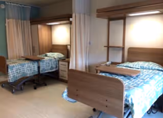 A room with two hospital-style beds, each with blue patterned bedding and wooden headboards and footboards. Each bed has an overbed table and a small bedside cabinet. There are privacy curtains between the beds and a light above each bed.