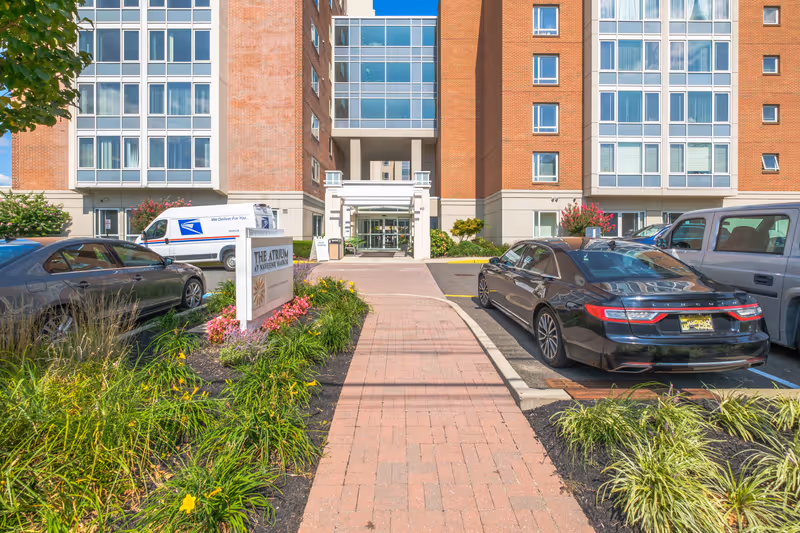 Front entrance of The Atrium at Navesink Harbor facility with a brick walkway leading to glass doors. The building is multi-story with large windows and brick exterior. There are parked cars on both sides of the walkway and landscaped flower beds with green plants and colorful flowers along the path.
