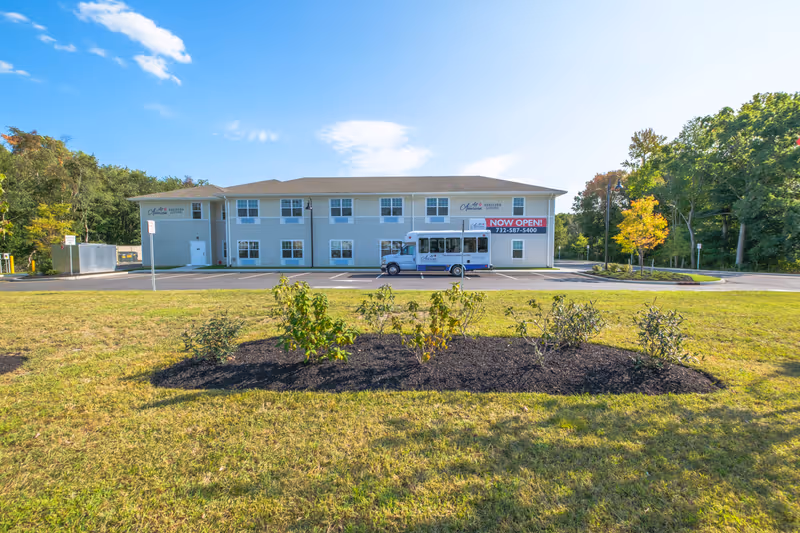 Exterior view of a two-story assisted living facility building with a parking lot in front. A small white shuttle bus is parked in the lot. There is a landscaped area with small bushes and grass in the foreground. The building has multiple windows and signs that read 'All American Assisted Living' and a large banner stating 'NOW OPEN! 732-587-5400'. Trees and a clear blue sky are visible in the background.