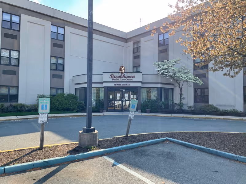 Exterior front view of Brookhaven Health Care Center, a multi-story building with large windows and an entrance canopy. There are two reserved parking signs and a tree with autumn leaves near the entrance.
